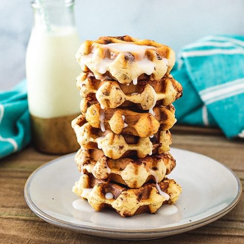 A stack of cinnamon roll waffles sitting on a plate with a cup of milk in the background